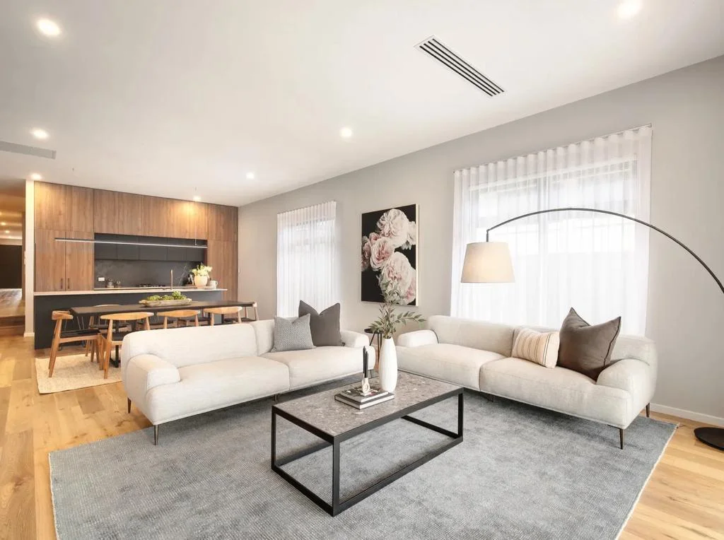 Modern living room with light-gray sofas, a dark rectangular coffee table on a gray rug, a floor lamp, and artwork on the wall. The open kitchen and dining area with wooden accents are in the background. Perfect for deep cleaning to maintain its pristine condition.