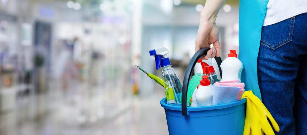 A person carries a blue bucket brimming with various cleaning supplies and yellow gloves in a brightly lit indoor setting, ready for one-off cleaning services.